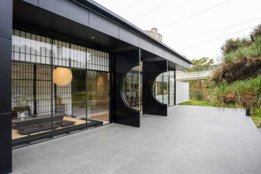 Çağdaş bir binanın taş kaldırımlı avlusu boyunca geniş açılı çekim, benzersiz dairesel kapılar ve sakin bir çay odasına bir bakış gösteriyor. Görüntü, uyumlu bir yaşam ve yenilikçi bir mimari tasarım sunuyor. Yüksek kaliteli gerçek fotoğrafçılık