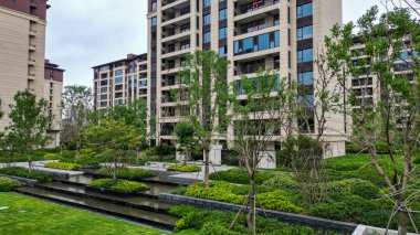 Şelaleler, taş verandalar ve apartmanlara karşı yemyeşil ağaçların bulunduğu sakin bir park manzarası. Görüntü lüks, meditasyon durgunluğu, doğal güzellik ve barışçıl toplum ortamını yansıtıyor. Yüksek kalite gerçek p