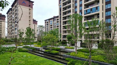 Çağdaş bir konut bahçesinin barışçıl manzarası katmanlı bir su özelliği, canlı çimenler ve apartmanlara karşı olgun ağaçlar. Görüntü mimari dengeyi, doğal uyumu ve huzur içinde yaşayan toplumu taşır. Yüksek kalite