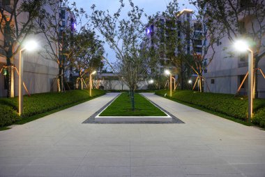 Alacakaranlık saatlerinde uzun, modern ışık direkleriyle aydınlatılan bahçedeki uzun, simetrik bir taş yol. Görüntü perspektif, geometrik düzen, akşam sessizliği ve manzara tasarımı sağlar. Sığ alan derinliğine sahip yüksek kaliteli gerçek fotoğrafçılık.