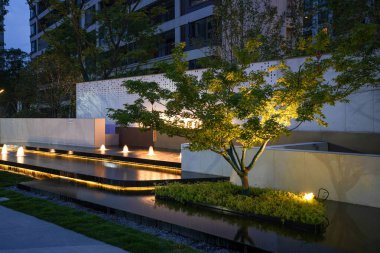 Aydınlatılmış mimari duvar ve ağaçların bitişiğindeki şaşaalı bir bahçe suyu manzarası. Görüntü karmaşıklığı, tasarım uyumu, meditasyonu ve akşam güzelliğini aktarıyor. Sığ alan derinliğine sahip yüksek kaliteli gerçek fotoğrafçılık