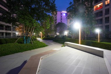 Parlayan direklerle aydınlatılan, geceleri ağaçlar ve çalılarla aydınlatılan bir yerleşim parkında huzurlu bir yol. Görüntü, akşam sükunetini, toplum yaşamını, mimariyi ve sükuneti aktarıyor. Yüksek kalitede gerçek fotoğrafçılık ve sığ fi derinliği