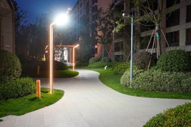 Parkta kıvrılarak ilerleyen zarif bir taş yürüyüş yolu, akşamları modern parlayan direklerle aydınlatılıyor. Görüntü, barışçıl hareket, manzara tasarımı, uyum ve yerleşim sükuneti taşır. Sığ alan derinliğine sahip yüksek kaliteli gerçek fotoğrafçılık.