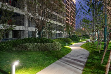 Geceleri sessiz bir konut parkında modern düşük seviyeli aydınlatma armatürleri tarafından aydınlatılmış temiz bir bahçe yolu. Görüntü huzur, güvenlik, modern peyzaj ve barışçıl bir atmosfer sağlıyor. Sığ alan derinliğine sahip yüksek kaliteli gerçek fotoğrafçılık.