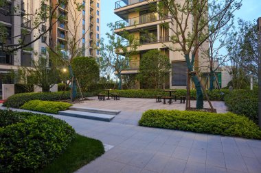 Alacakaranlıkta modern taş koltuklar ve ağaçların yer aldığı sakin, asfalt bir bahçe meydanı. Görüntü açık alan, toplum uyumu, huzurlu yaşam ve mimari tasarım taşır. Sığ derinliğe sahip yüksek kaliteli gerçek fotoğrafçılık