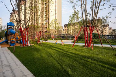 Çimenlikten geçerek modern bir oyun parkına doğru renkli ekipmanlarla çevrili, ağaçların çerçevelediği konut ortamında bir manzara. Görüntü neşe, toplum güvenliği, açık hava eğlencesi ve konut yeşilliği taşır. Sığ desenli yüksek kaliteli gerçek fotoğrafçılık