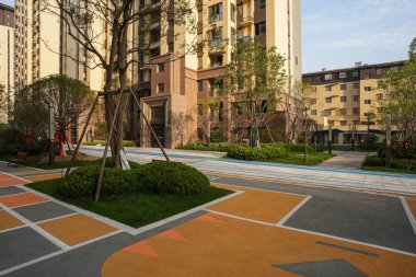 Geometrik renkli zemin yüzeyleri olan ve yemyeşil bir bahçe parkına entegre edilmiş modern bir yerleşim yeri manzarası. İmaj, toplumsal bağlılık, tasarım yeniliği, oyuncu atmosfer ve yerleşim uyumu taşır. Yüksek kaliteli gerçek fotoğraf.