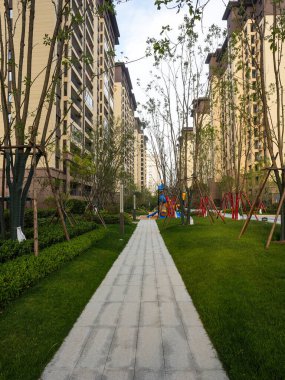 Temiz, yeşil bir parkın içinden geçen taştan bir patika. Uzakta bir çocuk oyun alanı var. Görüntü, güvenli bir mahalle hayatı, aile dostu tasarım, doğa ve sükunet sağlıyor. Sığ derinlikli yüksek kaliteli gerçek fotoğrafçılık