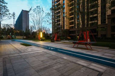 Geceleri sakin bir bahçe bahçesinde sıcak modern ışıklarla aydınlatılan, mavi bir faaliyet pisti olan temiz bir plaza manzarası. Resim, akşam keyfi, güvenlik, toplum ve huzurlu şehir yaşamını aktarıyor. Yüksek kaliteli gerçek fotoğrafçılık