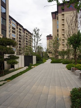 Modern apartmanlar ve parktaki gür ağaçlar arasında dönüp duran taştan bir plaza manzarası. Görüntü şehir ölçeğini, toplum açıklığını, dingin yeşilliği ve tasarım düzenini aktarıyor. Sığ alan derinliğine sahip yüksek kaliteli gerçek fotoğrafçılık.