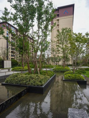 Yansıyan bir gölet, modern asfalt ve apartman mimarisine karşı yemyeşil ağaçların bulunduğu canlı bir bahçe. Görüntü dengeyi, doğa entegrasyonunu, lüksü ve toplumsal huzuru sağlıyor. Sığ derinlikli yüksek kaliteli gerçek fotoğrafçılık