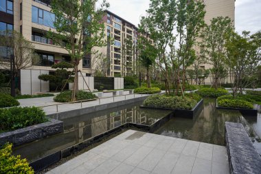 Temiz yansıyan göleti, taş kaldırımı ve yemyeşil ağaçları olan sakin, modern bir bahçe meydanı. Görüntü dinginliği, lüks yaşamı, tasarım dengesini ve huzur dolu bir yaşam ortamını aktarıyor. Sığ alan derinliğine sahip yüksek kaliteli gerçek fotoğrafçılık.