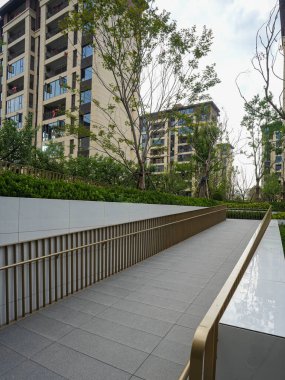 Minimalist metal tırabzanları olan şık, modern bir giriş rampası, yüksek ağaçlı, manzaralı bir yerleşim alanının içinden geçiyor. Görüntü evrensel tasarım, kapsayıcılık, mimari uyum ve barışçıl açık hava bağlantısını iletir. Yüksek kalite gerçek p