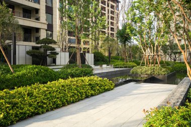 Çerçevesi yemyeşil yapraklar ve ağaçlarla çevrili sakin bir yansıtma havuzuna bitişik gevrek bir taş yolu bulunan sakin bir bahçe manzarası. Görüntü meditasyon dengesi, lüks açık hava tasarımı ve barışçıl şehir doğası taşır. sh ile yüksek kaliteli gerçek fotoğrafçılık