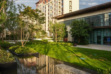 Açık bir yansıtma havuzunun, manikürlü çimlerin ve modern apartmanlara karşı olgun ağaçların bulunduğu güneşli bir bahçe parkı. Görüntü, huzurlu bir yaşam, mimari denge ve doğal uyumu temsil ediyor. Yüksek kalite gerçek fotoğraf.