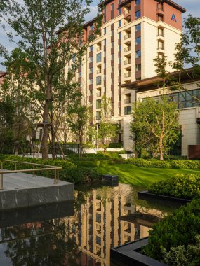 Bereketli yeşil peyzaj ve yerleşim alanındaki uzun ağaçlarla çevrili modern bir yansıtma havuzunun huzurlu manzarası. Görüntü lüks bir yaşam tarzı, toplum uyumu, mimari tasarım ve meditasyon sükuneti taşır. Yüksek kaliteli gerçek fotoğrafçılık