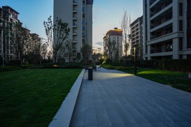 Huzurlu bir akşam manzarası, modern binalara doğru uzanan, yumuşak ışıklandırmayla kaplı bir yerleşim yeri yolu. Görüntü güvenliği, toplumsal ahengi, akşam sükunetini ve mimari güzelliği aktarıyor. Kaliteli gerçek fotoğrafçılık ve sığ bir derinlik.