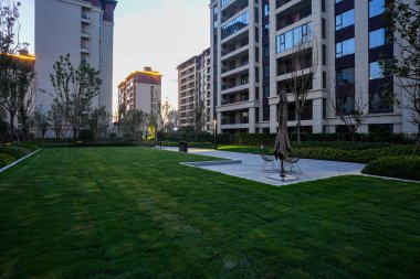 Akşamları, modern apartman yapılarıyla çevrili bir konut parkında geniş, manikürlü bir bahçe meydanı. Görüntü açıklık, toplum eğlencesi, sakin açık hava atmosferi ve lüks yaşam içeriyor. Sığ ve yüksek kaliteli gerçek fotoğrafçılık