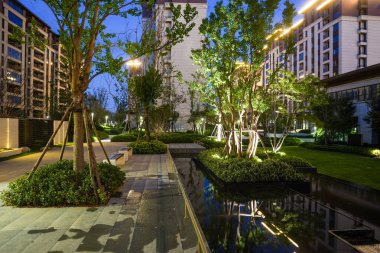 Modern bir bahçe parkının gece çekimi, yansıtan bir gölet, ağaçlar ve sıcak mimari aydınlatma. Görüntü, akşam sükuneti, lüks, toplum tasarımı ve sakin bir atmosfer sağlıyor. Sığ ve yüksek kaliteli gerçek fotoğrafçılık