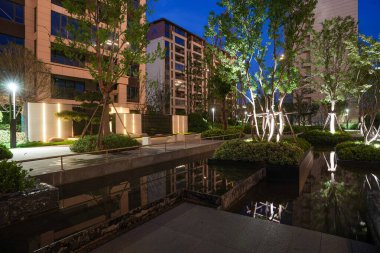 Günümüz mimarisini ve alacakaranlık altında yemyeşil ağaçları yansıtan sakin, aydınlık bir meskun su özelliği. Görüntü, lüks tasarımı, toplumsal durgunluk, meditasyon ve son model açık hava ortamını taşır. sh ile yüksek kaliteli gerçek fotoğrafçılık