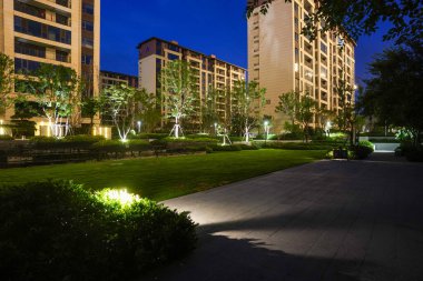 Temiz çizgili bahçe parkı, manikürlü çimenler, çalılar ve sıcak gece ışıkları, huzurlu bir şehir alanı sağlıyor. Görüntü tasarım düzeni, sükunet, doğa ve üst düzey yerleşim atmosferi taşır. Yüksek kaliteli gerçek fotoğrafçılık zekası.