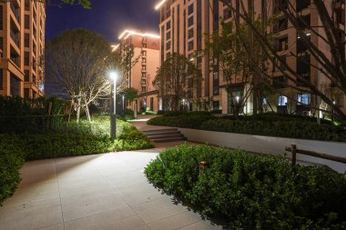 Modern zemin ışıklarıyla aydınlatılan ve huzurlu bir gece inzivası oluşturan bir yerleşim parkında dolambaçlı, parlak, temiz bir yol. Görüntü tasarım uyumu, akşam sakinliği, açık hava yaşam tarzı ve mimari güzelliği aktarıyor. Yüksek kaliteli gerçek fotoğrafçılık