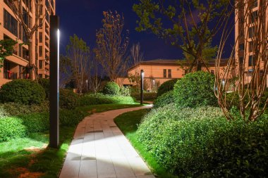 Modern ışık direkleriyle aydınlatılan, geceleri sakin bir yerleşim bölgesinde yemyeşil alanda süzülen berrak bir bahçe geçidi. Görüntü huzur, mimari tasarım, güvenlik ve huzur dolu ev yaşamını aktarıyor. Sığ ve yüksek kaliteli gerçek fotoğrafçılık