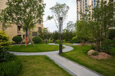 Ağaçlar ve modern mimari binalar tarafından çerçevelenmiş, peyzajlı bir konut parkında dolambaçlı, sakin, taş döşenmiş bir yürüyüş yolu. Görüntü şehir uyumu, akşam eğlencesi, yapısal tasarım ve toplumsal refahı iletir. Yüksek kaliteli gerçek fotoğrafçılık.