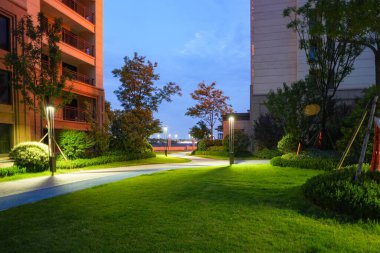 Alacakaranlıkta bir yerleşim parkında, manikürlü peyzaj ve ağaçlarla çevrili, parlak, temiz bir patika. Görüntü, mahalle barışı, güvenlik, mimari detay ve akşam sükunetini aktarıyor. Şerifle kaliteli gerçek fotoğrafçılık.