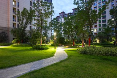 Modern direkler tarafından aydınlatılan, manikürlü çitlerle ve ağaçlarla dolu, canlı bir yerleşim parkında dolambaçlı, zarif bir taş geçit. Görüntü toplum huzurunu, tasarım dengesini, akşam hayatını ve doğal cazibesini aktarıyor. Yüksek kalite gerçek çekim.