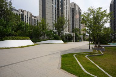 Temiz bir gökyüzü altında, yemyeşil çimlerle ve modern apartmanlarla çevrili taştan yapılmış bir yerleşim alanının geniş açılı görüntüsü. Görüntü açık toplum alanı, yapısal düzen, sağIık ve huzurlu günlük yaşam taşır. Yüksek kalite gerçek fotoğraf.