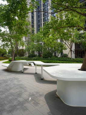 Bir ağacın etrafına inşa edilmiş modern beyaz bir bankın yakın çekimi, bir bahçede huzurlu ve yaratıcı bir oturma alanı sağlıyor. Görüntü mimari zarafet, açık hava konforu, refah ve toplum tasarımı taşır. Sığ ve yüksek kaliteli gerçek fotoğrafçılık