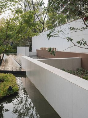 Bir su özelliğini çevreleyen, çevreleyen ağaçları ve ışığı yansıtan parlak, modern bir beyaz duvar. Görüntü minimalizm, saflık, mimari denge ve barışçıl şehir doğasını aktarıyor. Sığ alan derinliğine sahip yüksek kaliteli gerçek fotoğrafçılık.