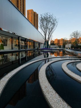 Açıklama: Modern mimari çizgilerin ve bir su özelliğine yansıyan dairesel manzara şekillerinin yer aldığı dinamik bir kompozisyon. Görüntü karmaşık tasarım, yapı ve sanatsal şehir planlaması içeriyor. Yüksek kaliteli gerçek fotoğrafçılık..