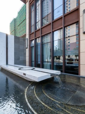 Cam bir binaya karşı taş ve dairesel detaylarla donatılmış minimalist bir su özelliği. Görüntü zen tasarımı, minimalizm ve mimari detay taşır. Yüksek kalite fotoğrafçılık.