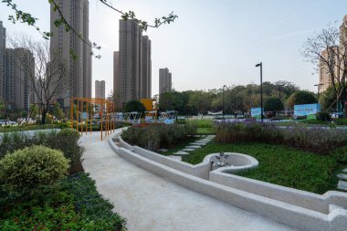 Tarif: Arka planda modern şehir yerleşim binaları olan, peyzajlı bir bahçede dolambaçlı beyaz bir patika. Görüntü, manzara dinginliği ve tasarım taşır. Yüksek kalite fotoğrafçılık.