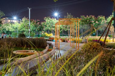 Gece huzurlu bir şehir parkı yolu, yumuşak altın ışıklarla aydınlatılmış ve yemyeşil bahçe yataklarıyla çevrili. Görüntü gece sükuneti, mimari ve tasarımı aktarır. Yüksek kalite fotoğrafçılık.