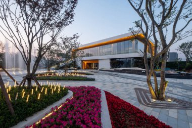 Alacakaranlıkta modern ışıklandırma, bina ve canlı bahçe yataklarının olduğu bir park alanı. Görüntü, gece bahçesinin tasarımını, yapısını ve kentsel güzelliğini yansıtıyor. Yüksek kalite fotoğrafçılık.