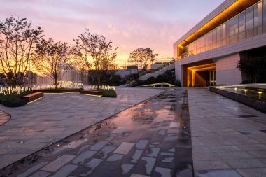 Tarif: Islak zemini, canlı günbatımı renklerini ve bina ışıklarını yansıtan modern bir plaza. Görüntü şehir havasını, mimari ayrıntıları ve tasarımı aktarır. Yüksek kalite fotoğrafçılık.