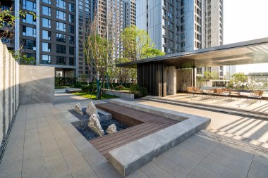 Açıklama: Bahçe taşları, ahşap süslemeler ve modern yapıların yer aldığı manzaralı bir park meydanı. Görüntü, barışçıl bir atmosfer ve açık hava tasarımı sağlıyor. Yüksek kalite fotoğrafçılık.