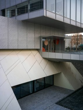 Çapraz mimari çizgilerin, taş kaplamaların, cam cephelerin ve yapılandırılmış batık avlunun bulunduğu modern bir bina kompleksine bakıyorum. Yenilikçi şehir tasarımı, kurumsal altyapı ve çağdaş yapı mühendisliği sunar..