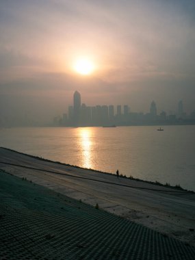 Geniş, sisli bir nehrin dikey görüntüsü. Altın sabah güneşi, eğimli bir taş setin yanındaki su yüzeyine mükemmel bir şekilde yansıyor. Huzurlu şehir doğasını, sabahın erken saatlerinde dinginliği ve huzurlu deniz manzarasını temsil eder..