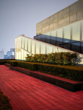 Kırmızı ahşap bir teras ve parlak pencereleri ve desenli metal bir ekranı olan çağdaş bir binanın dış cephesine uzanan yeşil bir çit içeren dikey kompozisyon. Modern mimari tasarım taşır.