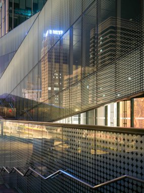 Süpürücü bir merdiven, aydınlatılmış cam cepheler ve geceleri yakınlardaki şehir binalarını yansıtan delinmiş bir metal ağ ekran içeren çağdaş mimari dış görünüş. Yenilikçi şehir tasarımı, ticari gayrimenkul ve modern kızılötesi