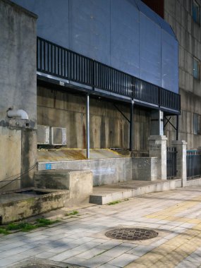 Metal tenteli ve klimalı eski bir beton bina duvarının yanında uzanan boş kaldırımlı yaya geçidi. Sahne, tarihi kentsel altyapı, şehir sokakları, kentsel çürüme ve sakin mahalle sokaklarını temsil ediyor.