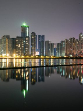 Aydınlatılmış yüksek modern gökdelenlerin dikey bileşimi geceleyin bir nehrin sakin karanlık yüzeyine mükemmel bir şekilde yansıyor. Sahne görkemli şehir manzaraları, metropol zenginliği, kentsel gayrimenkul ve barışçıl gece çevrecilerini temsil ediyor.
