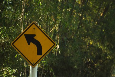 Yeşil ağaçlarla çevrili kırsal yol üzerindeki sarı sola uyarı işareti..