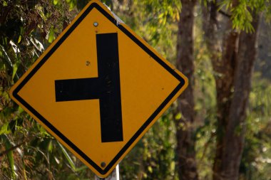 Sıcak gün ışığında ağaçlar ve doğal ormanlarla çevrili kırsal bir yol kenarında sarı yol kavşağı uyarısı..