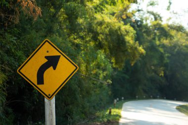 Çevresi yeşil ağaçlarla çevrili, dolambaçlı kırsal bir yolun yanındaki yol işareti. Sıcak, doğal gün ışığında..