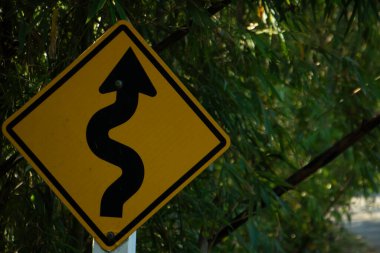 Yol kenarındaki sarı kıvrımlı yol uyarı levhası. Yeşil yapraklı doğal güneş ışığı..